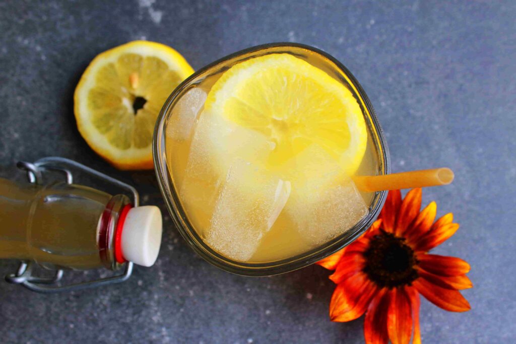 fermented lemonade in glass with ice and straw, top of a bottle with bottle stopper next to it, fresh lemon slice and flower