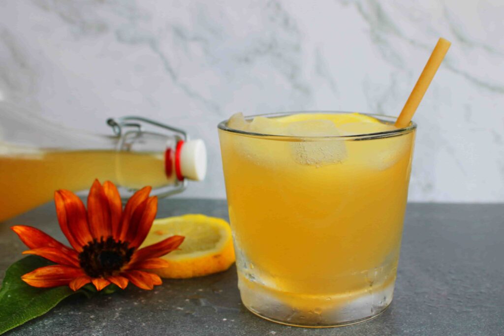 fizzy probiotic lemonade in glass with ice and straw, with fermented lemonade in bottle with stopper behind it