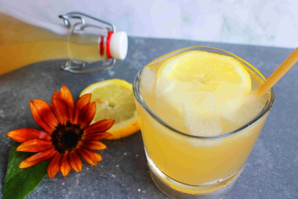fizzy probiotic lemonade in glass with ice and straw, with fermented lemonade in bottle with stopper behind it